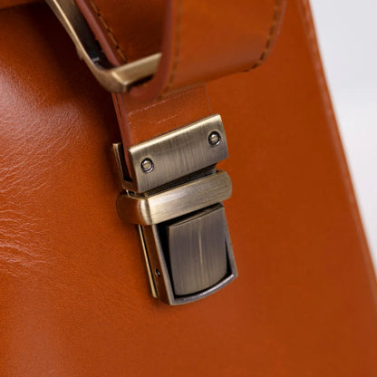 Close-up of a brown leather bag with a brushed brass buckle and strap detail against a white background.