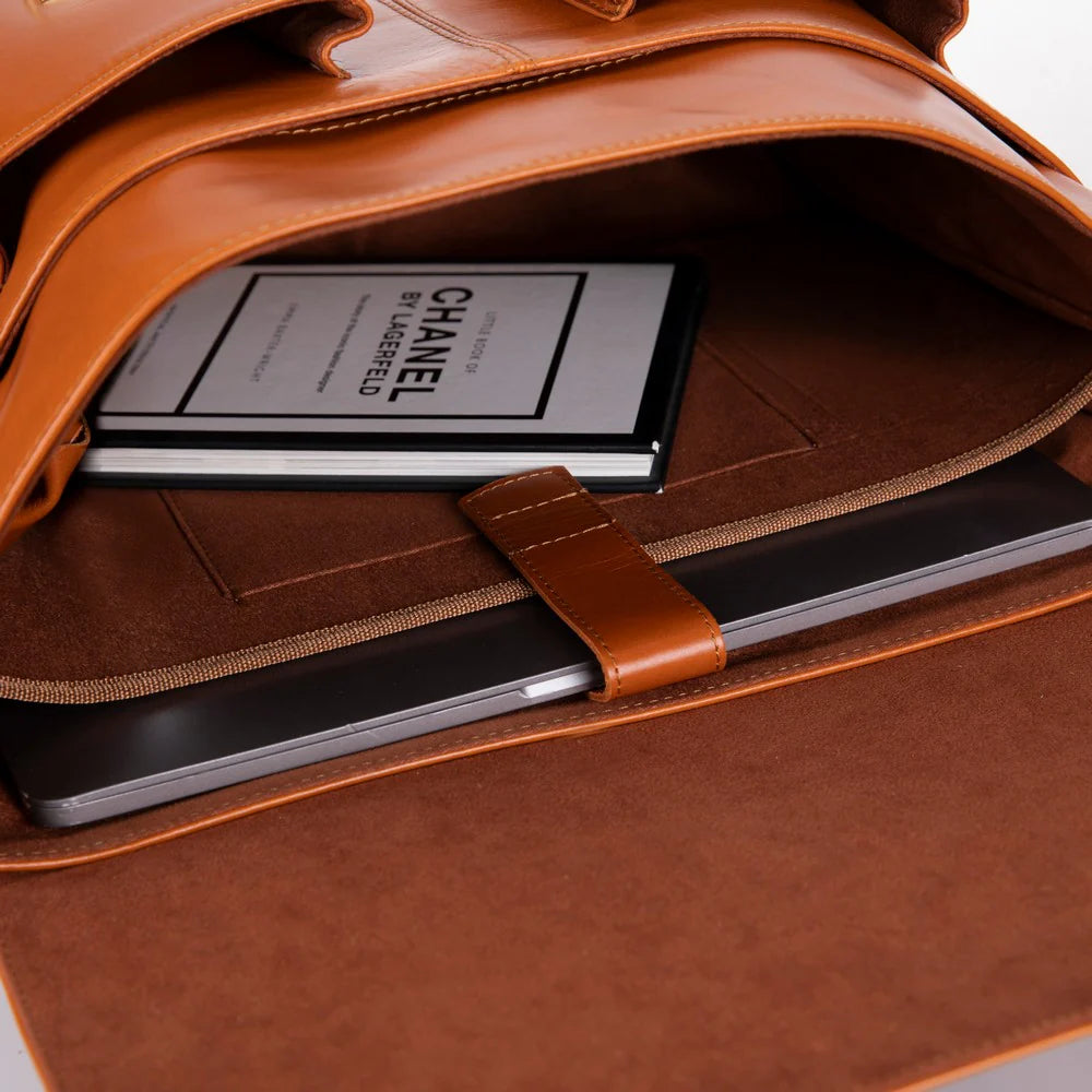 Close-up of a brown leather laptop sleeve or briefcase interior with a strap closure, holding a black tablet and a book with visible Chanel text.