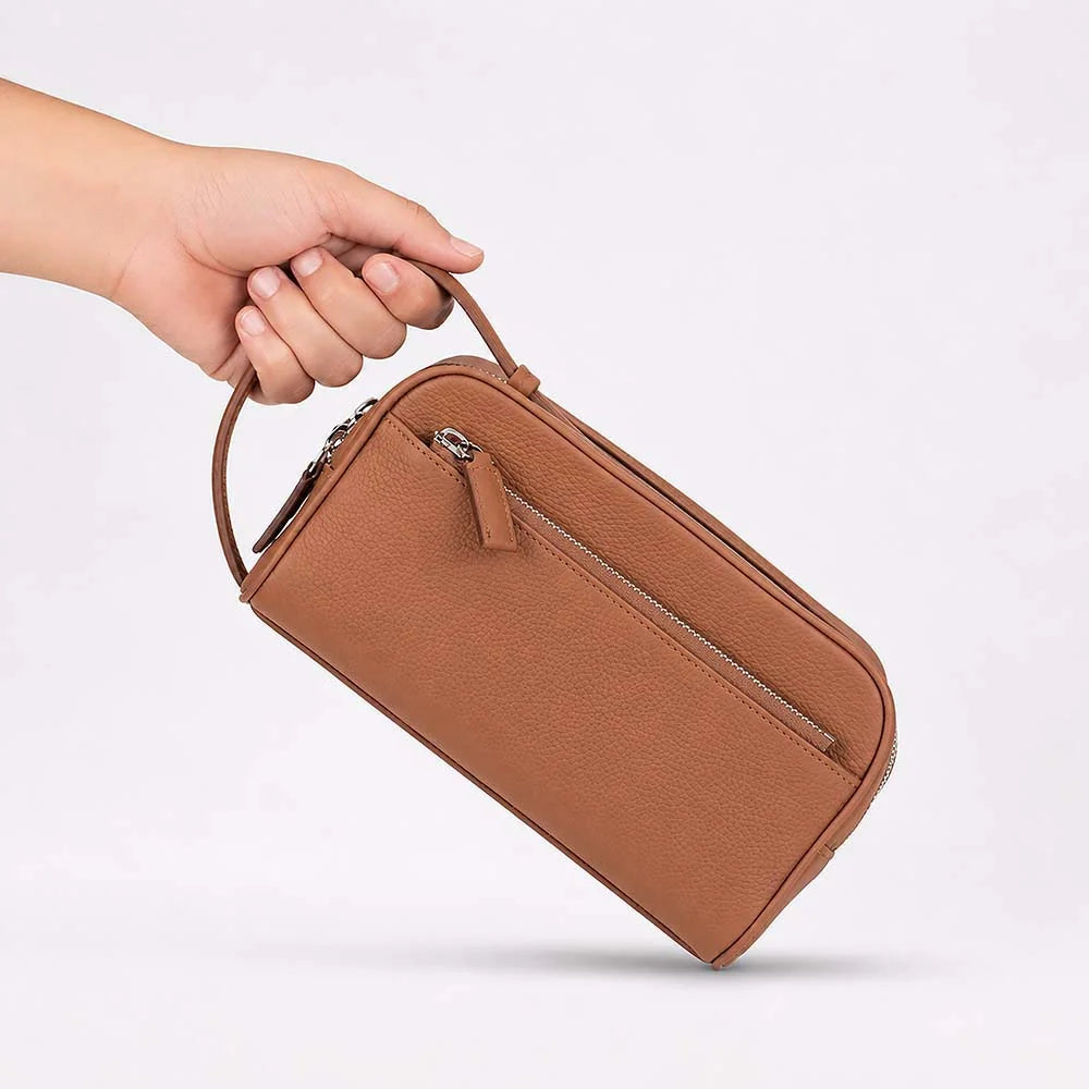 A hand holding a brown pebbled faux-leather zippered travel pouch with a top handle, shown against a clean white background.