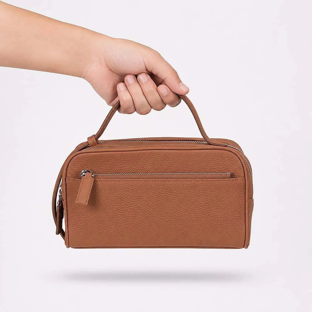 A brown pebbled leather top-handle bag being held by a hand against a clean white background.